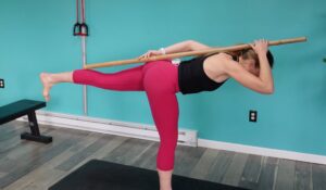 Dr. Chloe hinged forward at the hips with her left leg lifted holding a dowel to her spine as a hip hinge exercise also known as a single leg rdl